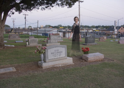 A lone figure stands somberly in a cemetery at dusk.