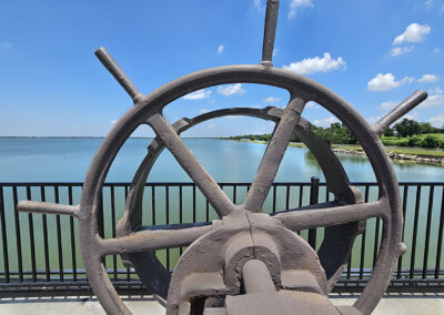 Rusty ship wheel against a blue sky and water backdrop.
