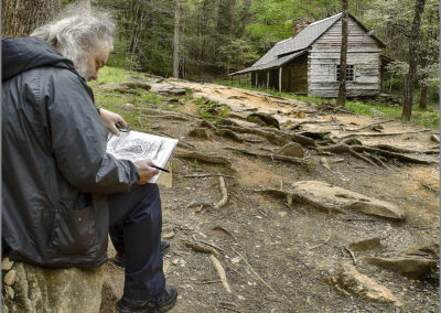 An elderly man reads a book while sitting outdoors near a rustic log cabin.