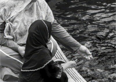 A child and an adult feeding fish by the water's edge.