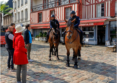 Two mounted police officers interact with pedestrians on a cobblestone street.