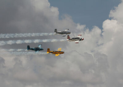 Five vintage airplanes flying in formation with smoke trails against a cloudy sky.