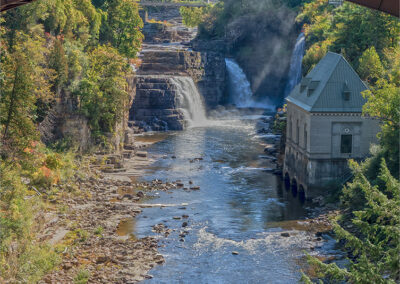 View of a river with waterfalls under a bridge and a building on the riverbank.
