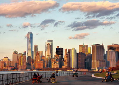People relax along a waterfront promenade with a city skyline at sunset.