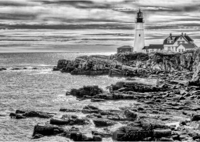 Black and white photo of a lighthouse on rocky shore under cloudy sky.