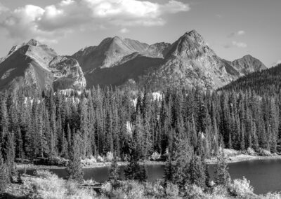 Black and white photo of a mountain range with a forest and lake in the foreground.