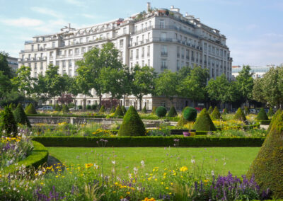 Elegant white building behind a vibrant garden with trimmed bushes and colorful flowers.