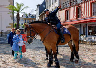 Mounted police officer gestures while talking to a woman on a cobblestone street.