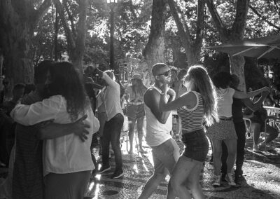 Couples dance closely in a sunlit park with tall trees.