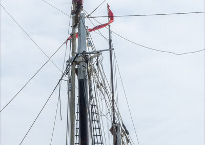 Person working atop a tall ship mast with rigging and a red flag.
