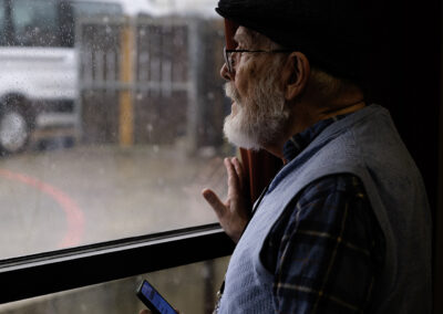 An elderly man with a beard looking out a rainy window while holding a phone.